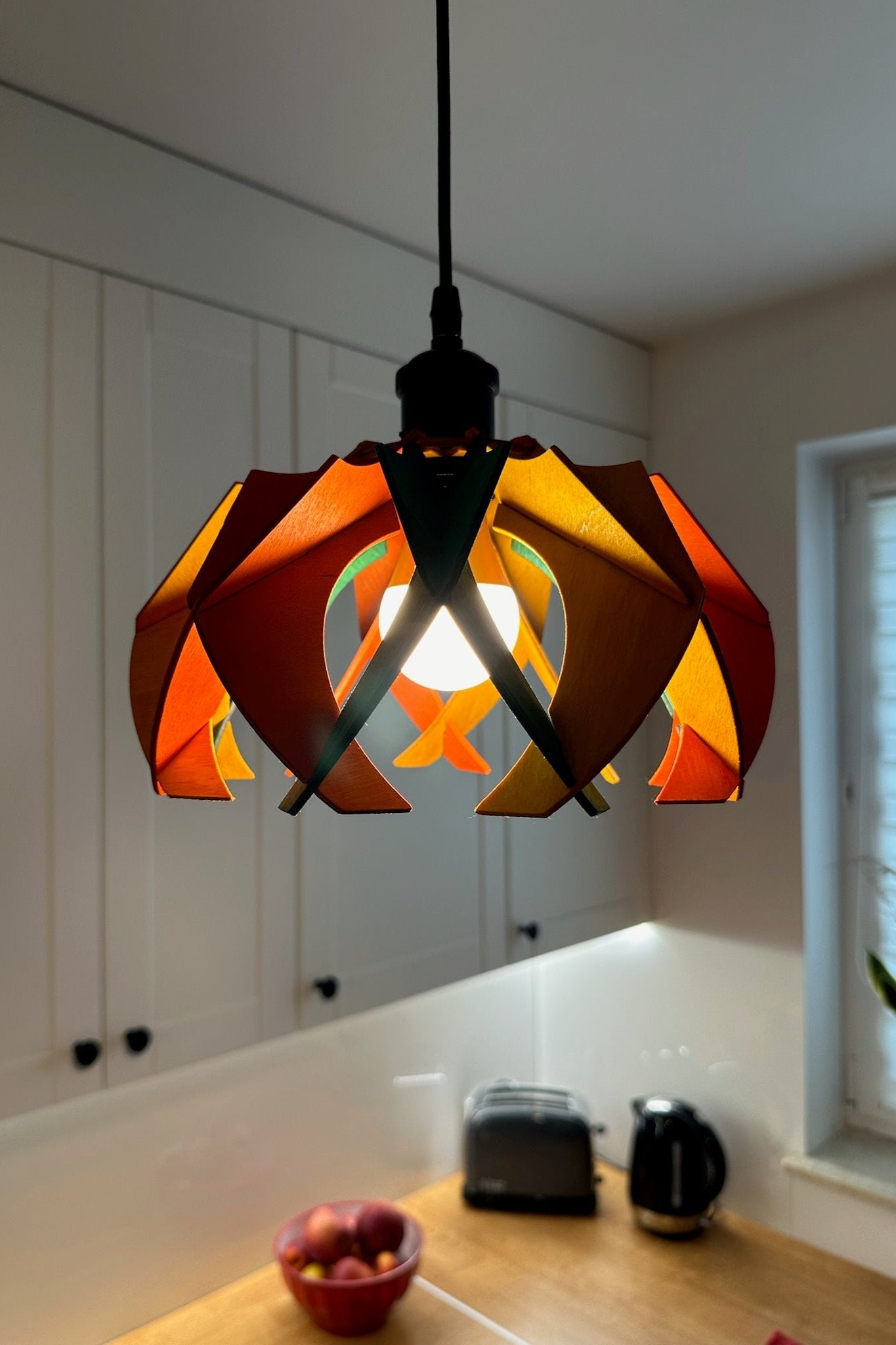 Small colorful pendant lamp in a kitchen setting, featuring a vibrant mix of green, yellow ochre, and scarlet stain. The lamp's petal-like wooden ribs create a dynamic and playful ambiance, with the light filtering through the vibrant colors.