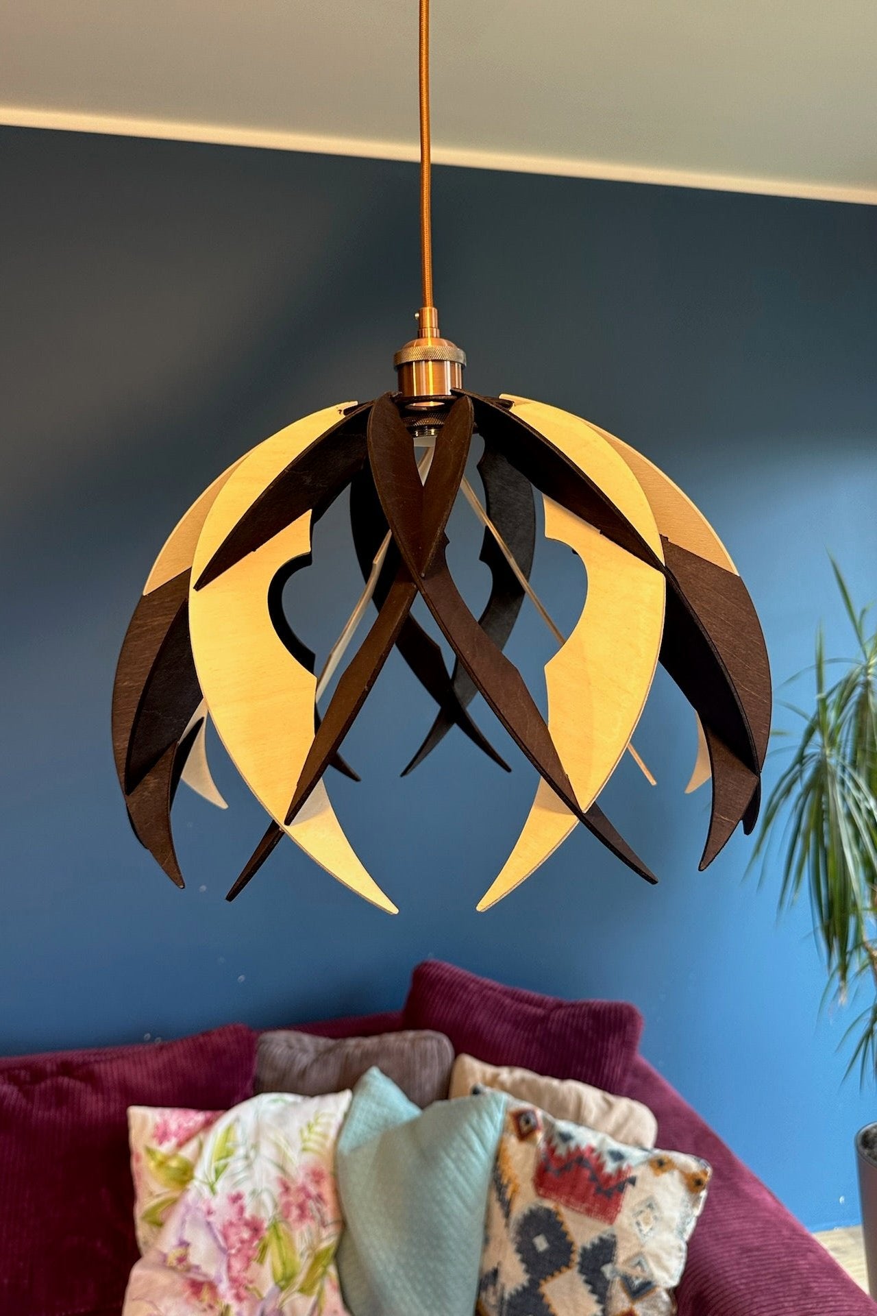 A black and white wooden pendant light with a dome-like shape, featuring pointed, curved ribs and circular cutouts. The light fixture hangs over a colorful sofa in a modern living room, with a copper cord and socket adding an industrial touch.