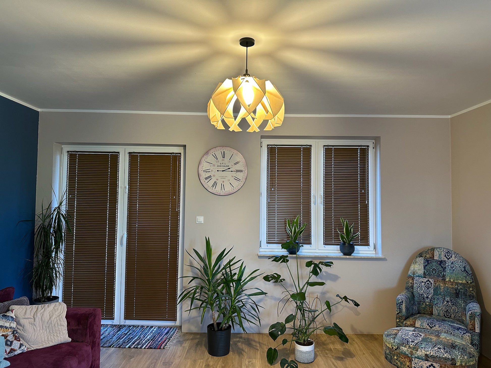 The large wooden pendant lamp hangs from the ceiling, illuminating a living room with warm, diffused light. The lamp&#39;s modern, flower-like design casts intricate light patterns on the walls and ceiling.