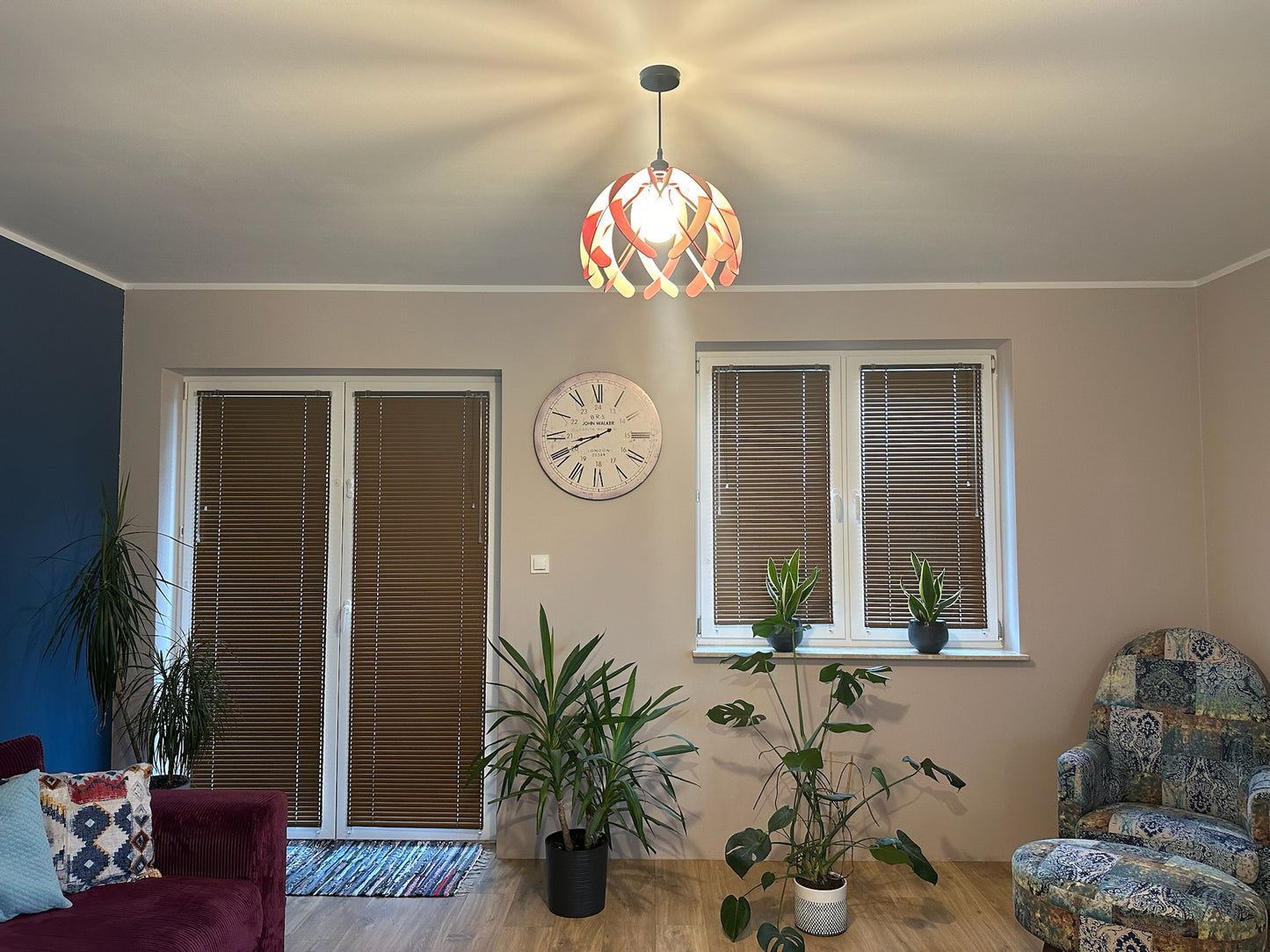 Pink and white wooden pendant lampshade hanging in a minimalist living room with green plants and modern furniture. The soft light creates a warm ambiance, and the black cord adds a sleek touch to the space.