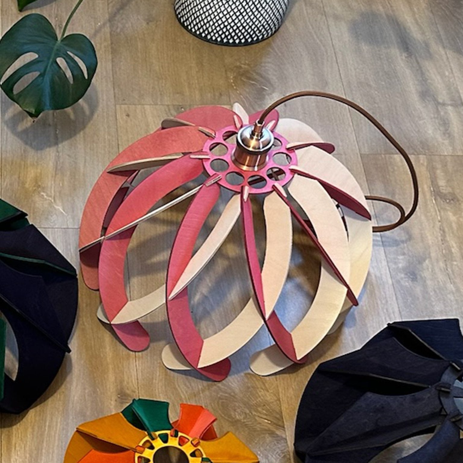 Pink and white wooden pendant lampshade placed on a wooden floor, alongside other color options such as black, green, and yellow. The lampshade is attached to a copper socket with a brown textile cord, offering a variety of customization choices.