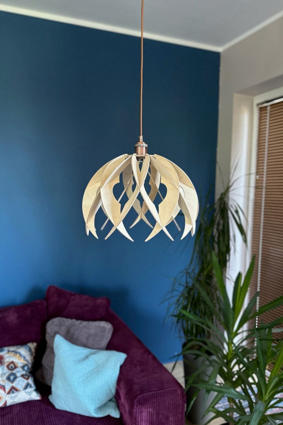 A dome-like wooden pendant light with a natural finish, featuring pointed, curved ribs and circular cutouts, suspended over a purple sofa with a teal accent pillow. The copper cord adds a warm touch to the modern, minimalist design.